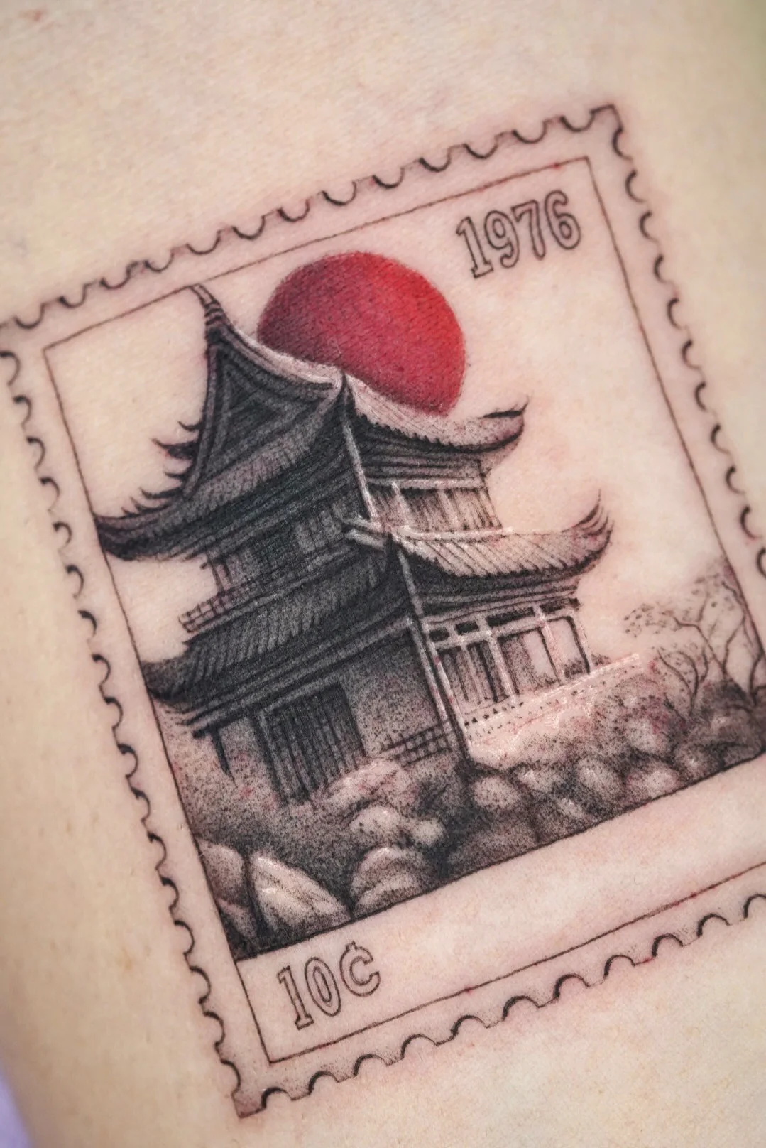 Postage stamp tattoo of a temple with a red sun, dated 1976